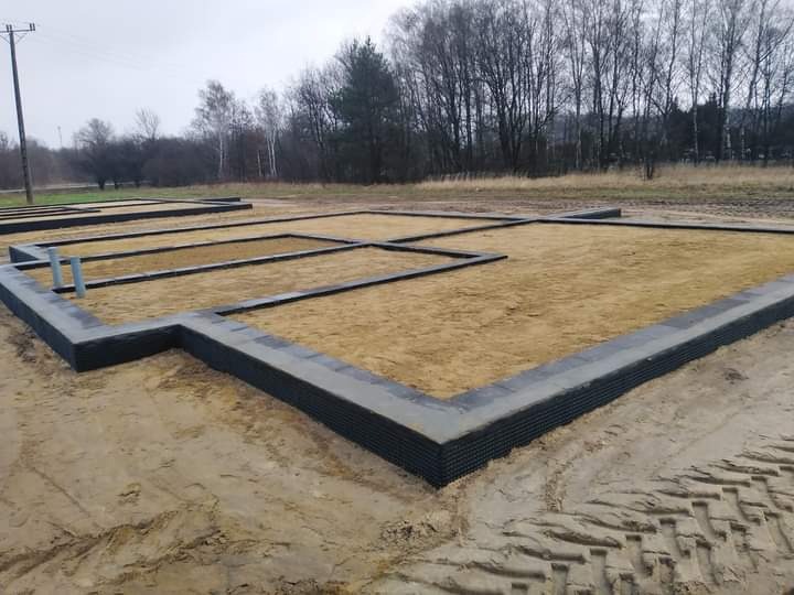 Przygotowany fundament pod budowę domu jednorodzinnego, widoczne szalunki wypełnione piaskiem oraz wystające rury kanalizacyjne.