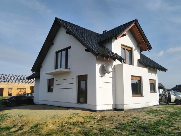 Nowoczesny dom jednorodzinny z białą elewacją i ciemną dachówką, widoczne okna z brązowymi ramami, antena satelitarna na ścianie, w tle budowa innego domu w stanie surowym otwartym.