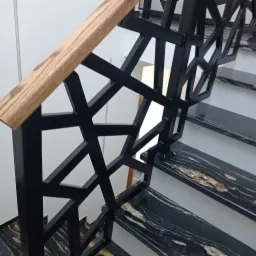 Nowoczesne schody z czarnymi, marmurkowymi stopniami i geometryczną, czarną balustradą z drewnianym pochwytem.