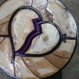 Okrągły, drewniany blat stolika kawowego z motywem pękniętego serca wypełnionym fioletową żywicą epoksydową i czarnymi metalowymi zdobieniami.