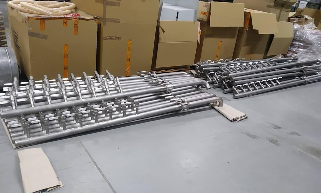 Stalowe rury z zaworami ułożone na szarym podłożu, w tle kartony, prawdopodobnie elementy instalacji przemysłowej.