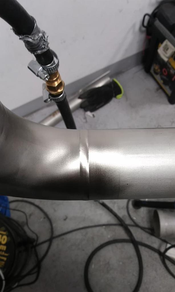 Spoina na rurze ze stali nierdzewnej, widoczny przewód doprowadzający gaz osłonowy z reduktorem, w tle narzędzia i sprzęt spawalniczy.