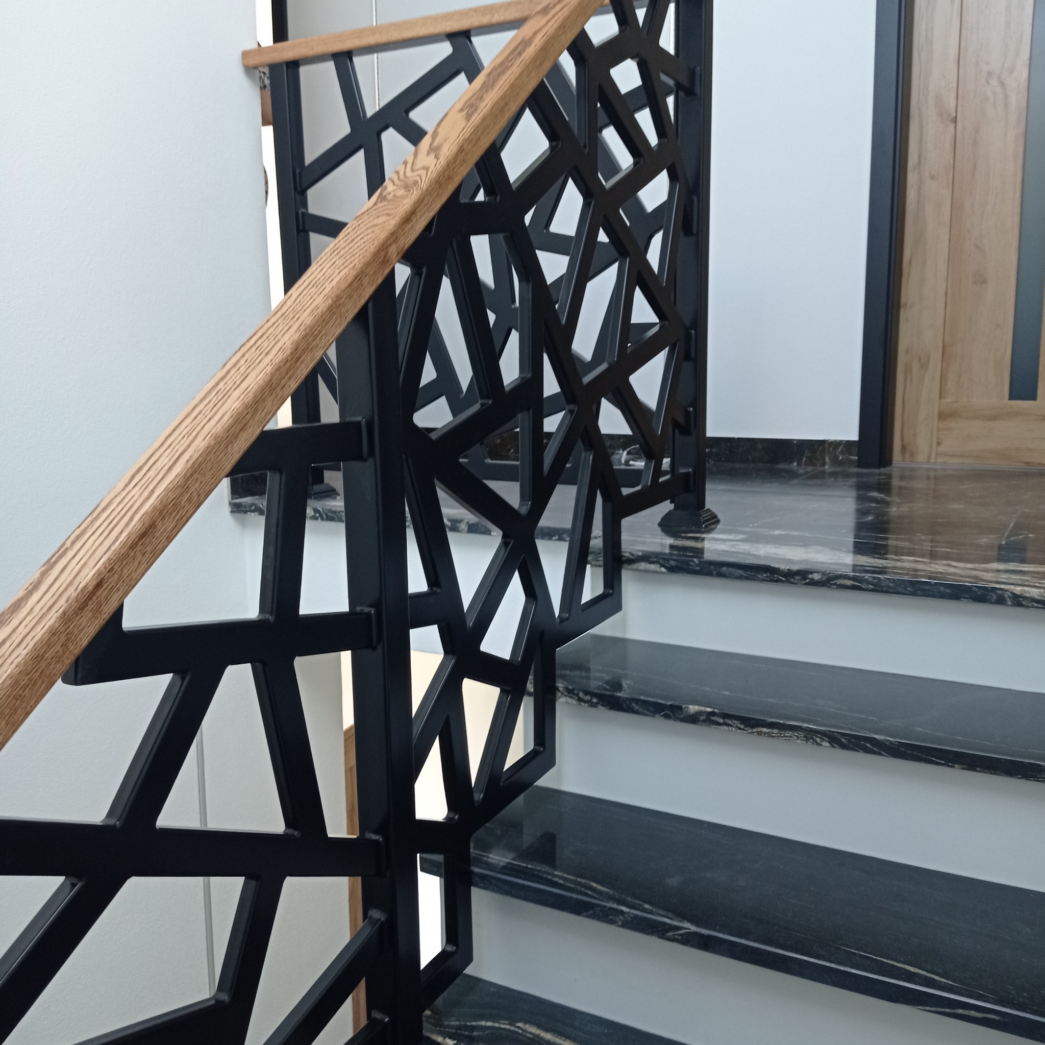 Nowoczesna balustrada schodowa z czarnego metalu o geometrycznym wzorze i drewnianym pochwytem, zamontowana przy schodach z czarnego marmuru.