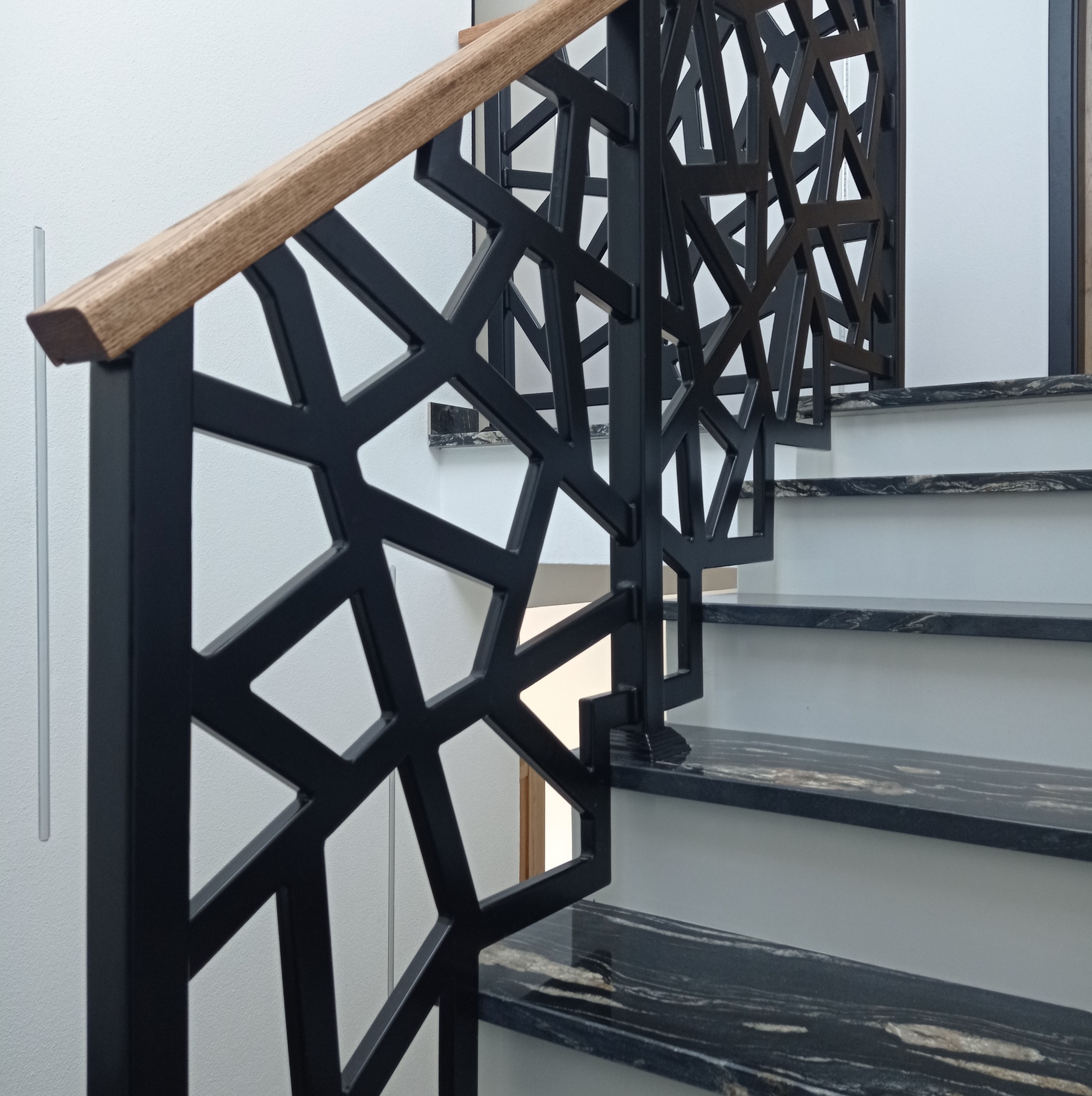 Nowoczesne schody z czarnymi, geometrycznymi poręczami metalowymi i drewnianym uchwytem, wykończone granitem w odcieniach szarości i czerni.