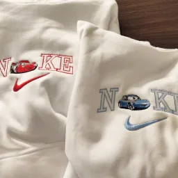 Dwa białe, bawełniane swetry z haftowanymi aplikacjami: czerwony samochód wyścigowy i logo Nike w kolorze czerwonym na jednym oraz niebieski samochód i logo Nike w kolorze niebieskim na drugim.