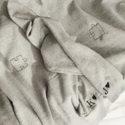 Dwie szare bluzy dresowe z motywem puzzli i inicjałami 'K' i 'J' z czarnym sercem, leżące na białym tle.