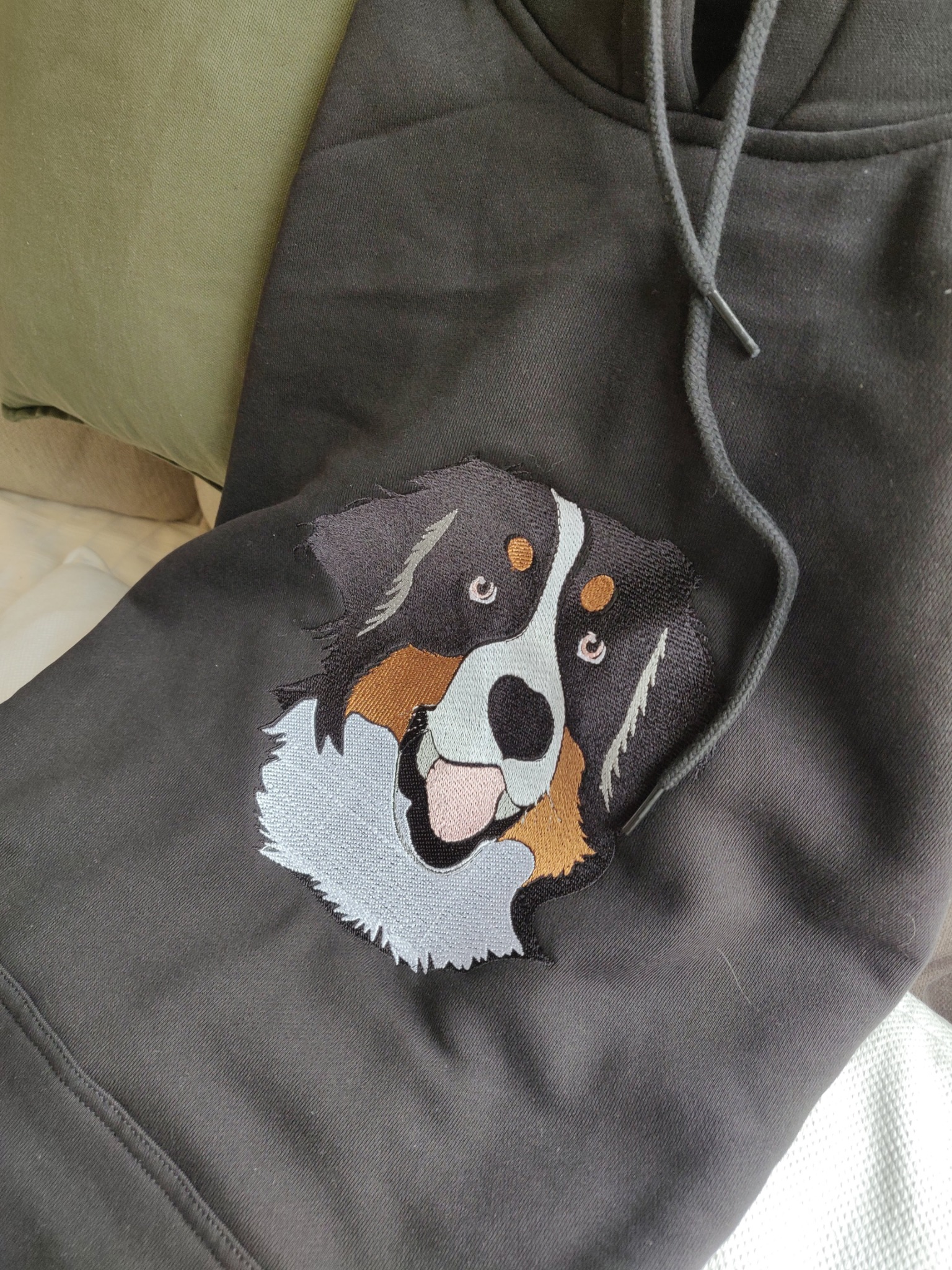 Ciemnoszara bluza z kapturem, na której wyhaftowano głowę psa rasy berneński pies pasterski z detalami w kolorach czarnym, brązowym i białym, leżąca na jasnej powierzchni obok zielonej poduszki.