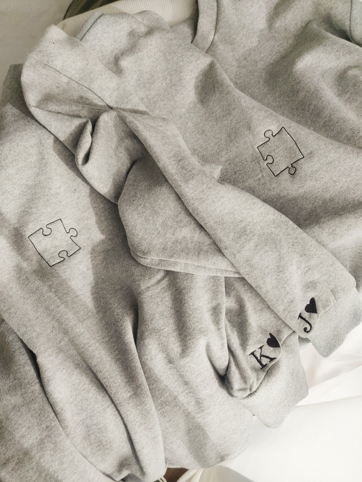 Dwie szare bluzy dresowe z motywem puzzli i inicjałami 'K' i 'J' z czarnym sercem, leżące na białym tle.