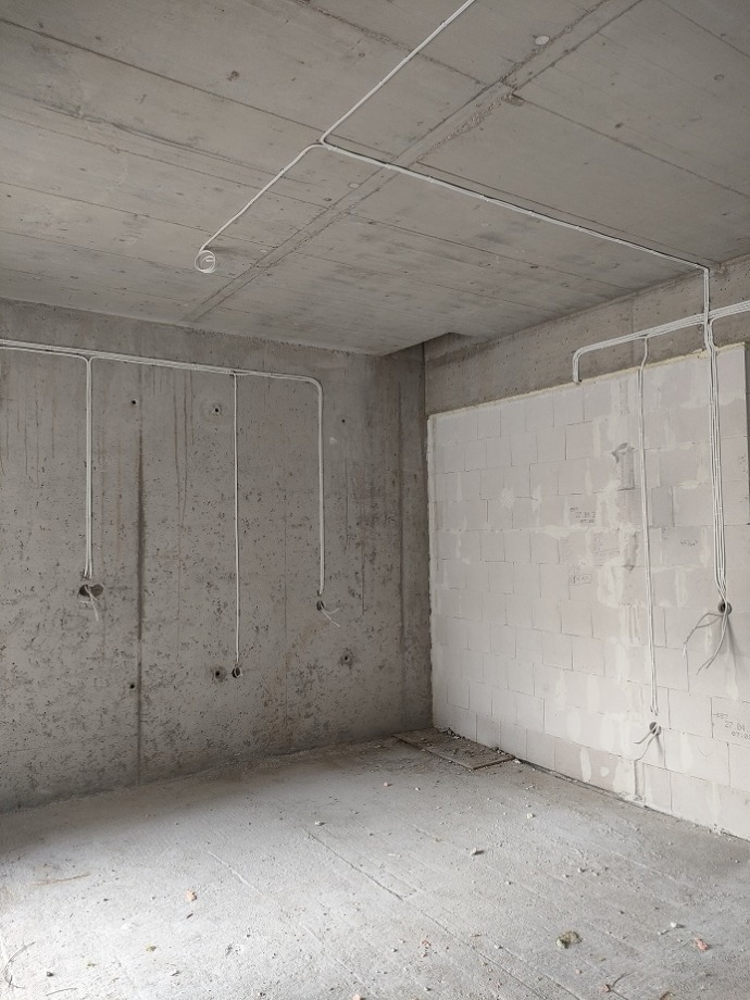 Surowy betonowy pokój w trakcie budowy z instalacją elektryczną poprowadzoną po ścianach i suficie, widoczne otwory na gniazdka.