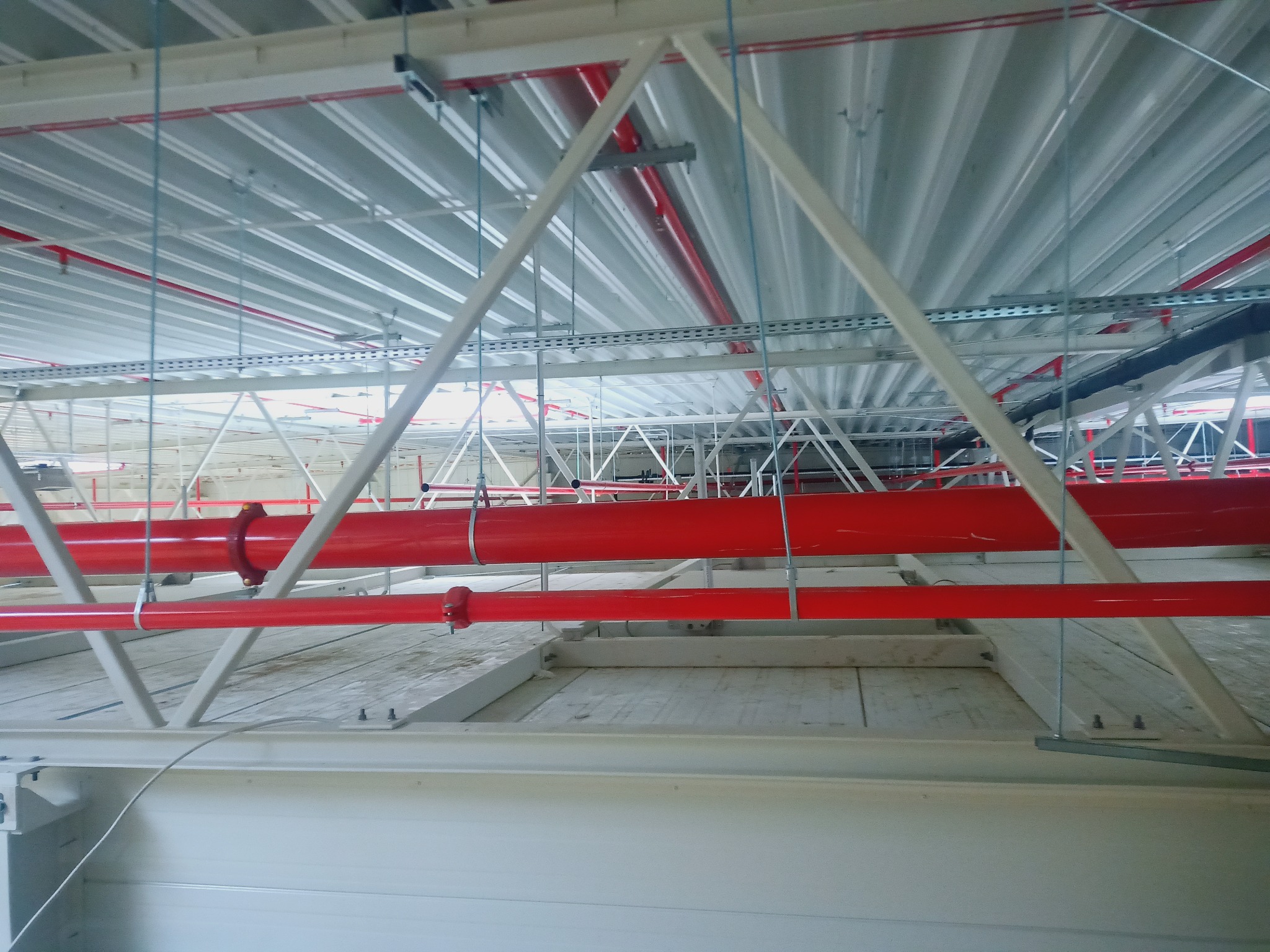 Wnętrze budynku w trakcie budowy z widoczną stalową konstrukcją dachu, czerwonymi rurami instalacji przeciwpożarowej oraz podwieszonymi elementami instalacji elektrycznej.