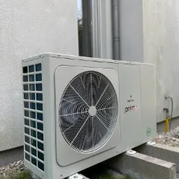 Zewnętrzna jednostka pompy ciepła Galmet Prima R32 zamontowana na betonowych wspornikach przy ścianie budynku z widoczną instalacją.