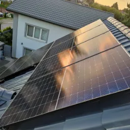 Panele fotowoltaiczne na grafitowym dachu domu jednorodzinnego, odbijające zachodzące słońce w tle zielone drzewa i fragment białej elewacji.