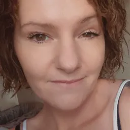 Portret kobiety z kręconymi włosami i delikatnym uśmiechem, wykonany w Białymstoku, z widocznymi ramiączkami koszulki na tle nieostrego wnętrza.