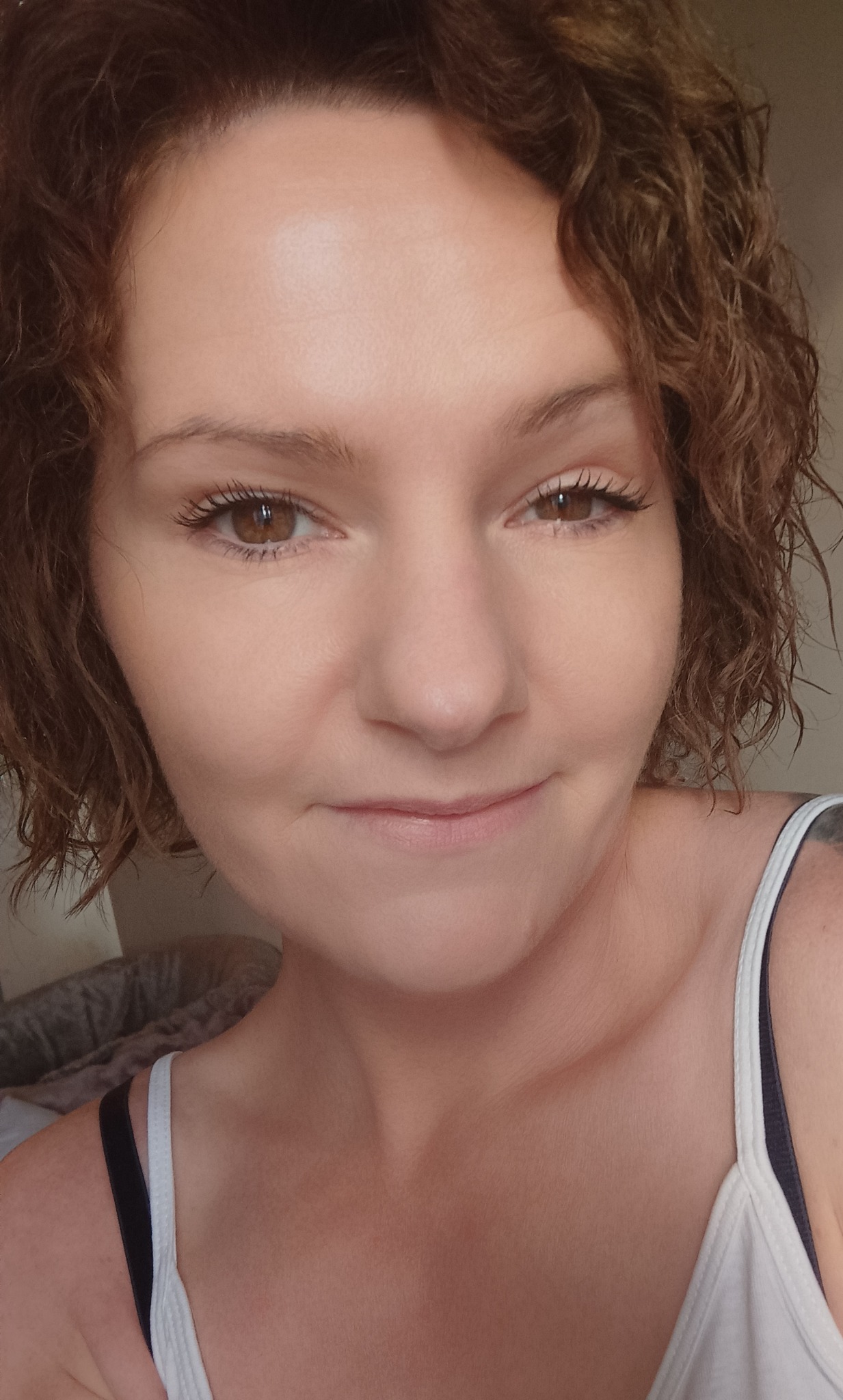 Portret kobiety z kręconymi włosami i delikatnym uśmiechem, wykonany w Białymstoku, z widocznymi ramiączkami koszulki na tle nieostrego wnętrza.