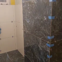 Układanie płytek imitujących marmur w łazience, widoczne niebieskie narożniki zabezpieczające oraz krzyżyki dystansowe.