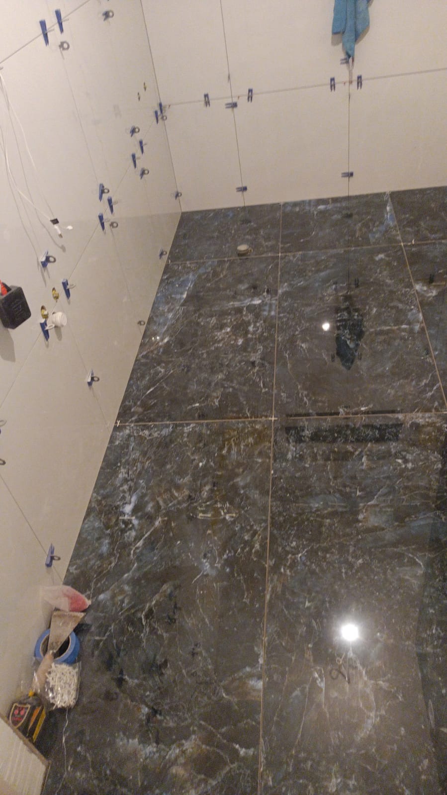 Fragment łazienki w trakcie układania płytek: jasne płytki na ścianach z niebieskimi klipsami poziomującymi i ciemne, błyszczące płytki imitujące marmur na podłodze, widoczne narzędzia i materiały...