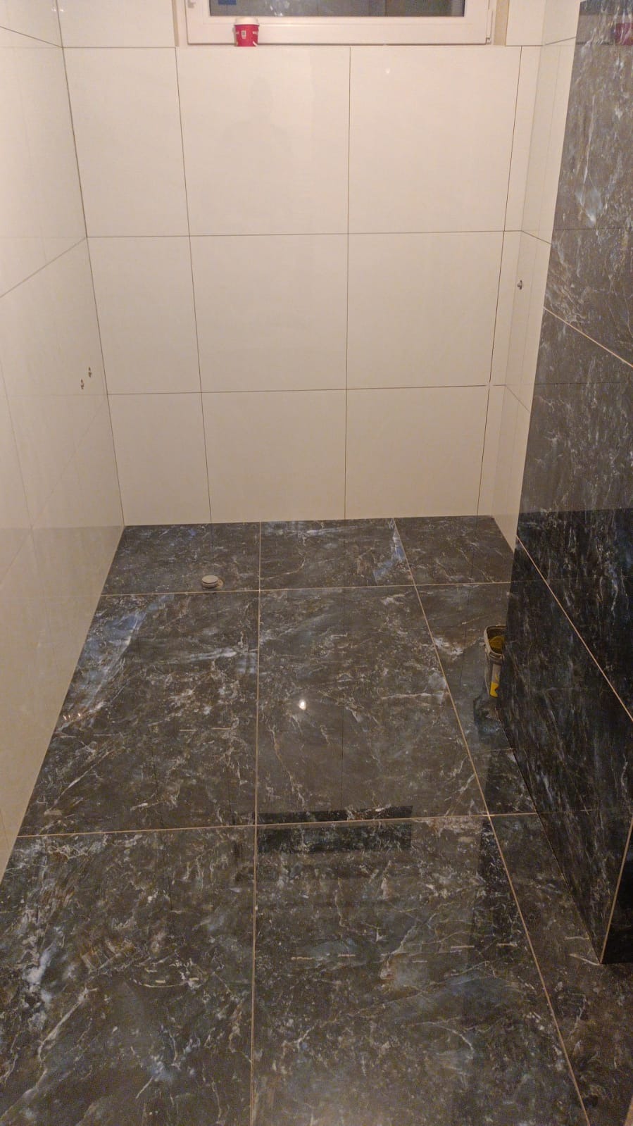 Wykończona łazienka z ciemnymi, marmurkowymi płytkami podłogowymi i jasnymi, kwadratowymi płytkami ściennymi, widoczny odpływ liniowy i wiaderko z narzędziami w rogu.