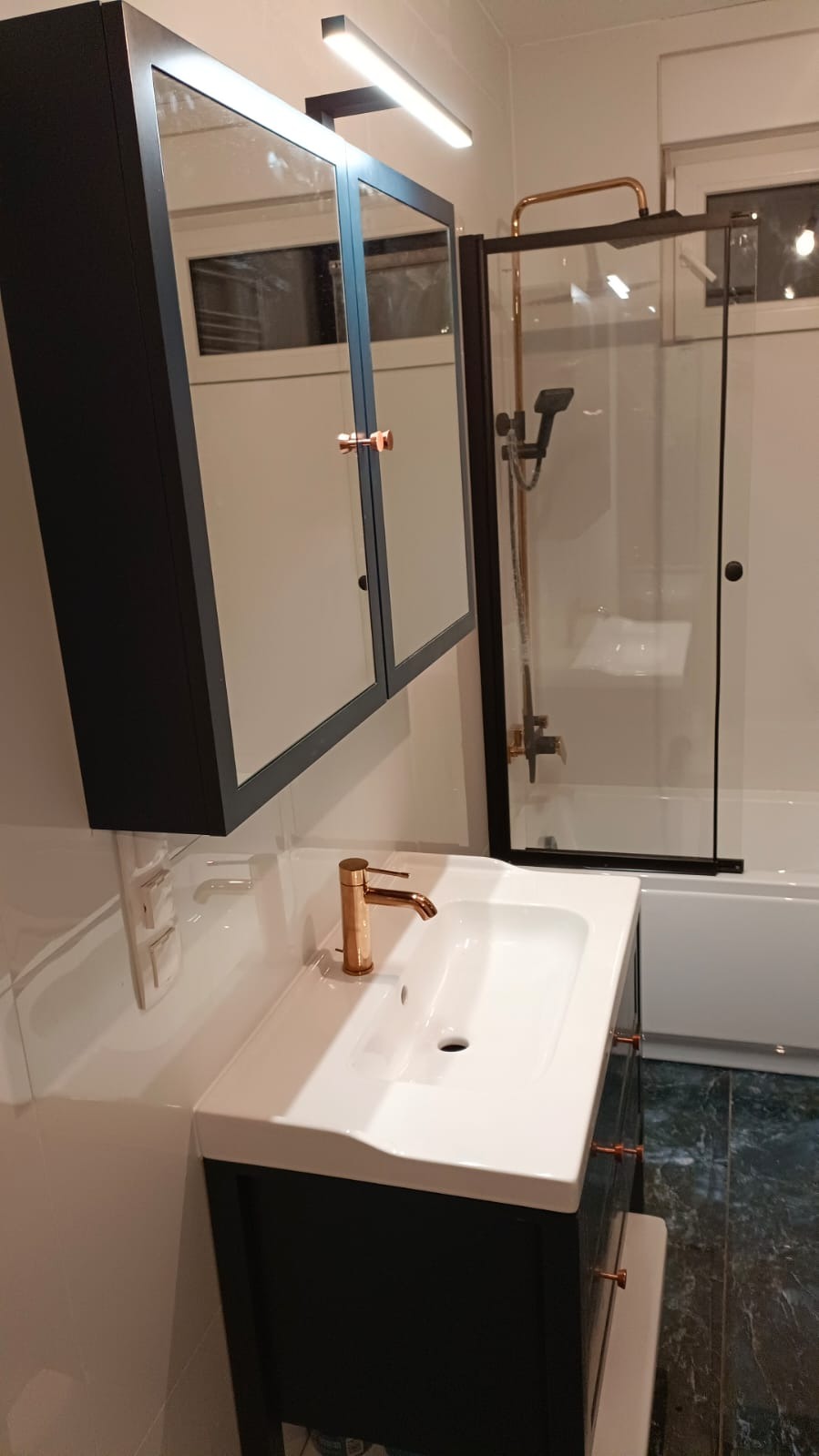 Nowoczesna łazienka z umywalką nablatową, szafką w kolorze ciemnego granatu z miedzianymi uchwytami, baterią w kolorze miedzi i kabiną prysznicową z czarnymi ramami.