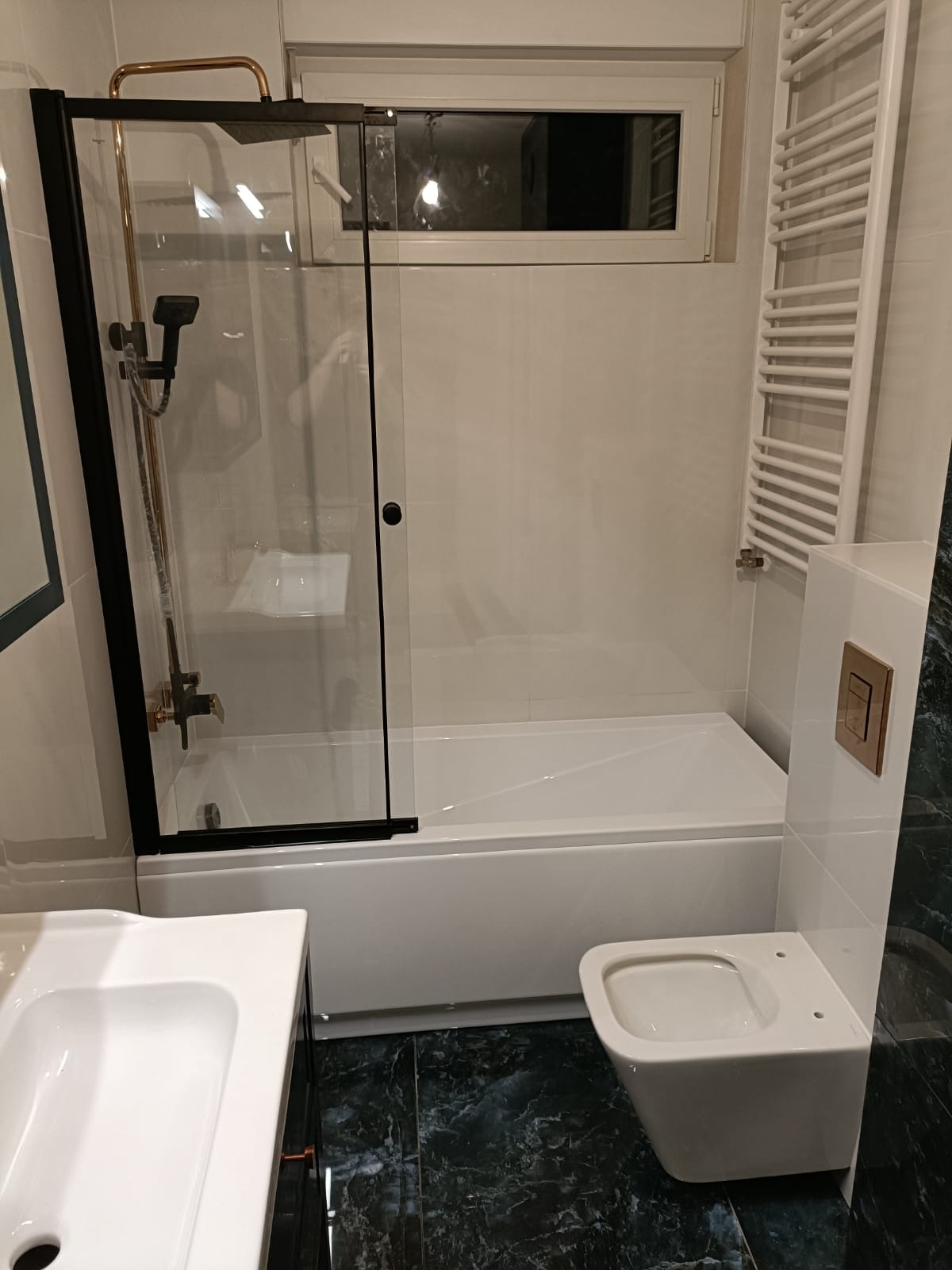 Wykończona łazienka z białą wanną z czarną ramą kabiny prysznicowej, wiszącą toaletą z przyciskiem w kolorze mosiądzu i grzejnikiem drabinkowym na ścianie z jasnych płytek, podłoga z płytek...