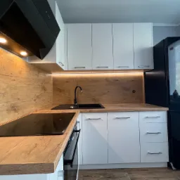 Nowoczesna kuchnia z białymi szafkami i blatem imitującym drewno, czarny zlew i bateria, podświetlenie LED nad blatem, czarny okap i lodówka.