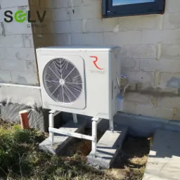 Biała jednostka zewnętrzna z wentylatorem marki Rotenso Aquami, zamontowana na metalowym stelażu na betonowych płytach przed budynkiem z bloczków betonowych.