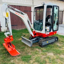 Mini koparka Takeuchi TB216 z łyżką hydrauliczną na trawie, przed budynkiem z cegły i drewnianymi drzwiami garażowymi.