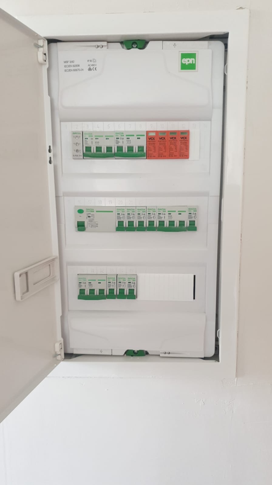 Otwarta, biała skrzynka elektryczna z bezpiecznikami i wyłącznikami, zamontowana w ścianie. Widoczne oznaczenia i logo 'epn'.
