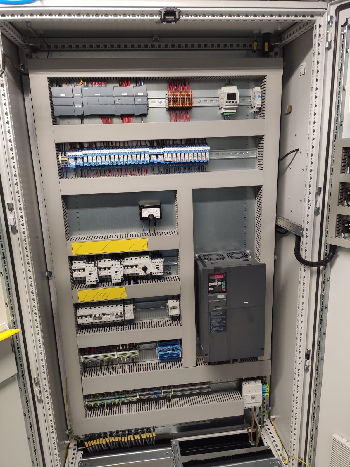 Rozdzielnica elektryczna w metalowej obudowie z widocznymi elementami sterującymi, przewodami i zabezpieczeniami. Układ schludny, zorganizowany wewnątrz szafy sterowniczej.