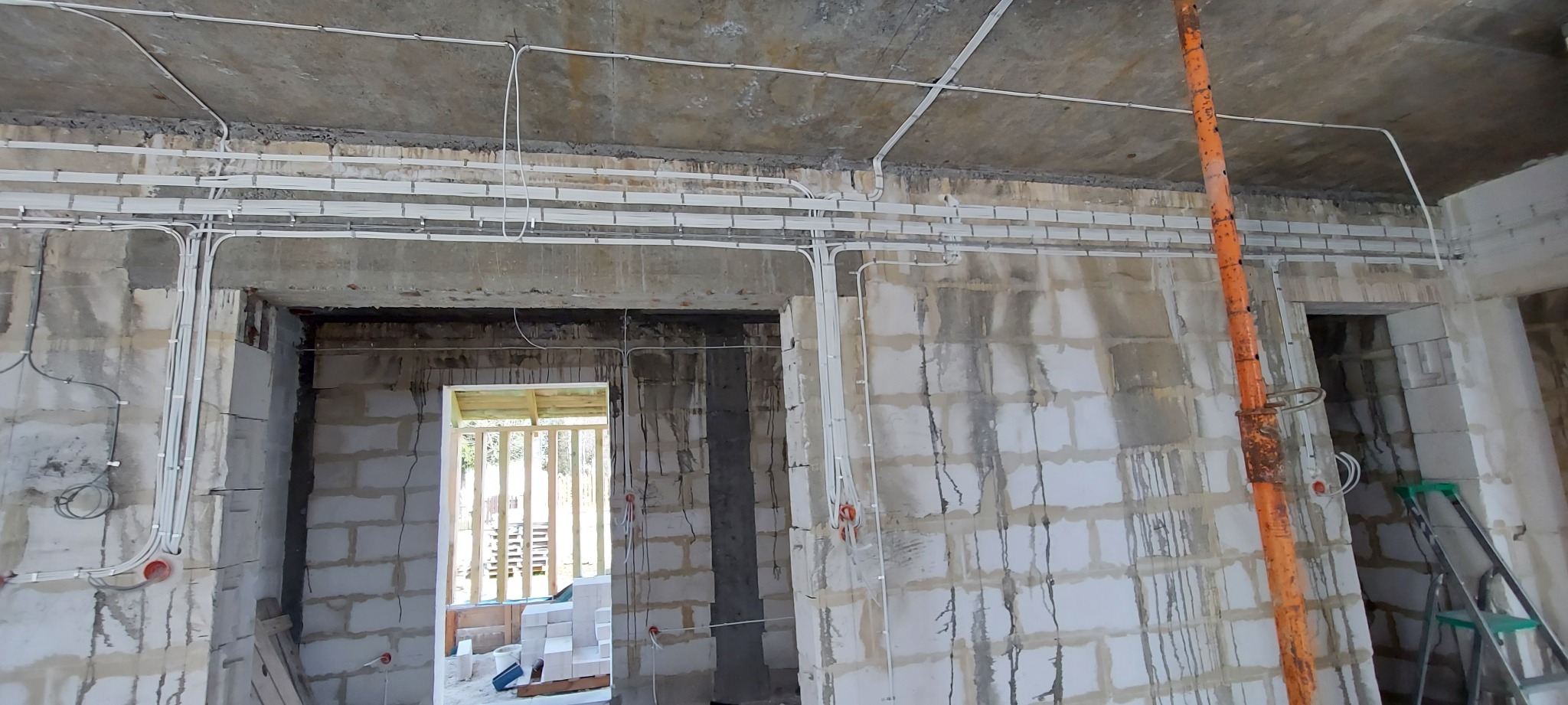 Wnętrze w stanie surowym z widoczną instalacją elektryczną natynkową, kable w białych listwach, puszki podtynkowe w ścianach z betonu komórkowego, podpora budowlana i drabina w tle.