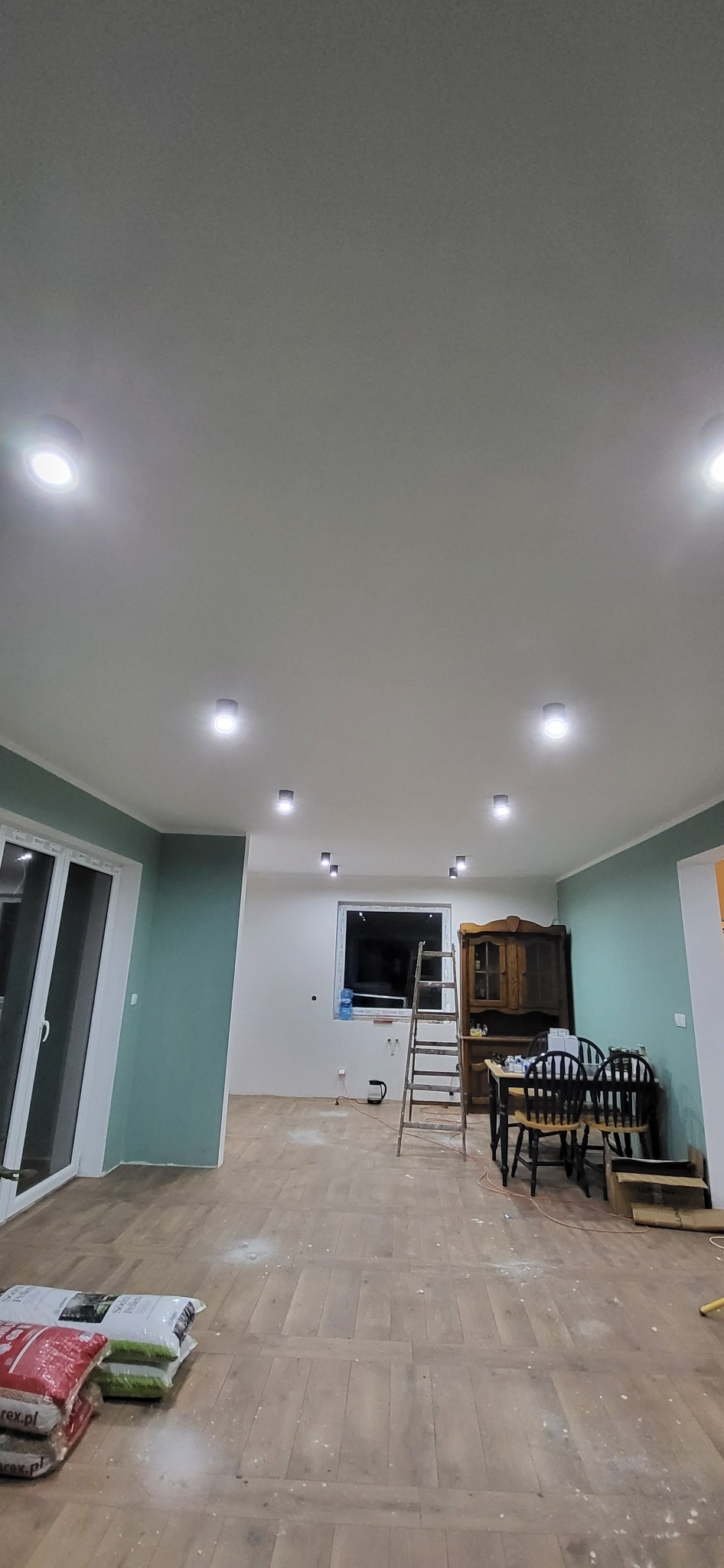 Wnętrze pokoju z zamontowanymi, okrągłymi lampami sufitowymi, drabina oparta o ścianę przy oknie, worki z materiałami budowlanymi na podłodze.