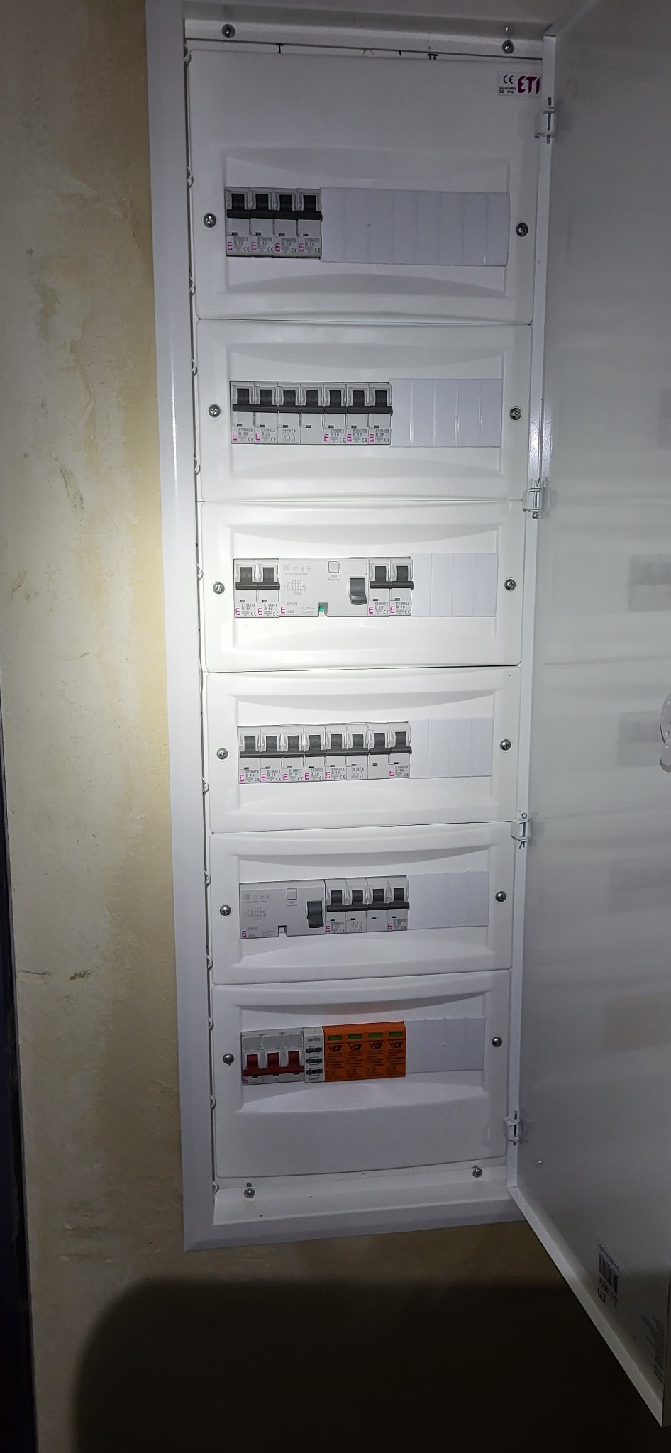 Otwarta, biała skrzynka elektryczna z zainstalowanymi bezpiecznikami i wyłącznikami różnicowoprądowymi, zamontowana na ścianie z widocznymi śladami malowania.