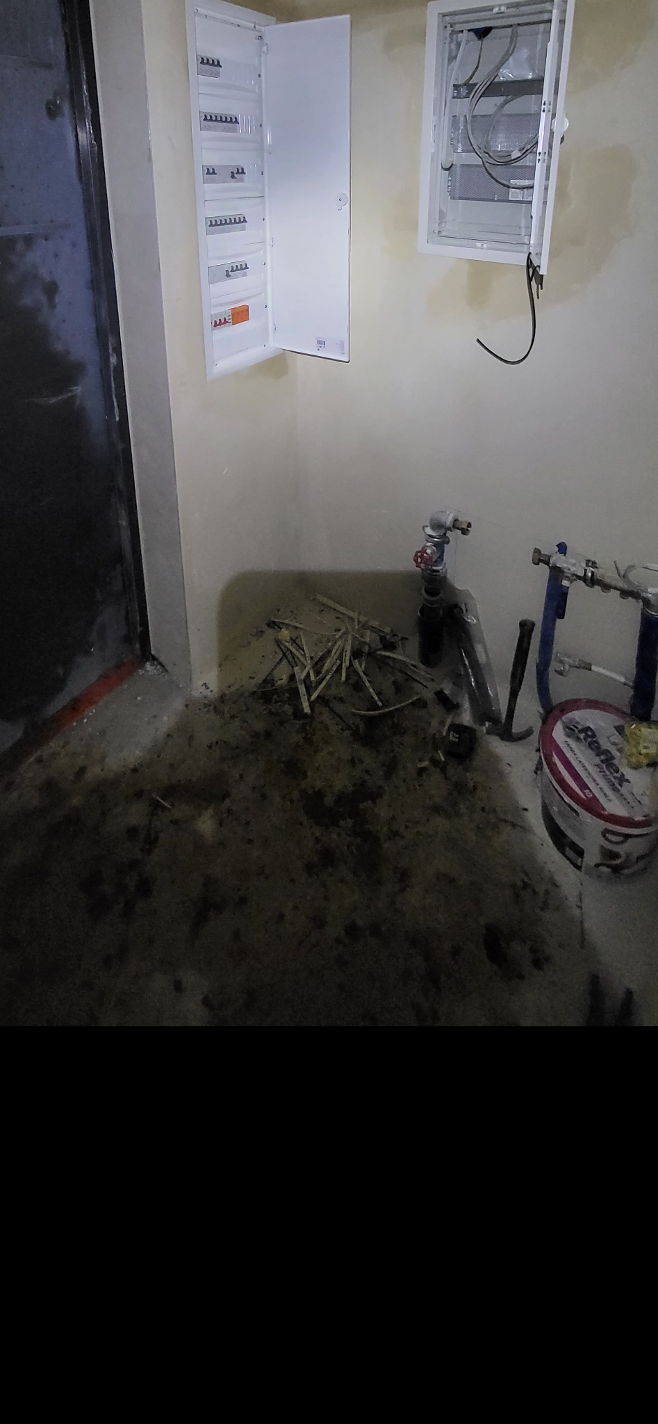 Dwie białe skrzynki elektryczne na ścianie, jedna otwarta z bezpiecznikami, druga pusta z kablami, na podłodze bałagan po remoncie, rury hydrauliczne i wiadro z farbą.