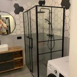 Nowoczesna łazienka z kabiną prysznicową typu walk-in z czarnymi profilami, heksagonalne płytki w odcieniach bieli i czerni na ścianie, drewniana szafka pod umywalką, okrągłe lustro z odbiciem...