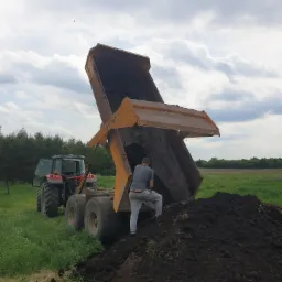 Ciężarówka z otwartą skrzynią wysypuje ziemię na pole; w tle widoczny czerwony traktor i pracownik w szarej koszulce.