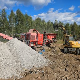 IMPERIAL FOREST - Czerwona kruszarka Terex Finlay J-960 i żółta koparka na placu budowy z hałdą gruzu na pierwszym planie, w tle drzewa i błękitne niebo z chmurami.
