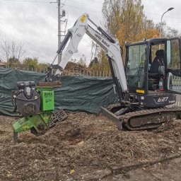IMPERIAL FOREST - Mini koparka Bobcat E27 z frezarką do pni Iwex, usuwająca pień drzewa. Wokół rozsypane wióry, w tle zielona plandeka i jesienne drzewa.