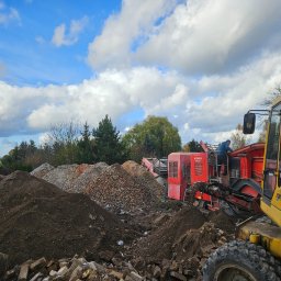 IMPERIAL FOREST - Żółta koparka i czerwona kruszarka do gruzu na placu budowy, z hałdami ziemi i gruzu. Operator na maszynie. Błękitne niebo z chmurami w tle. Widok na teren po rozbiórce.