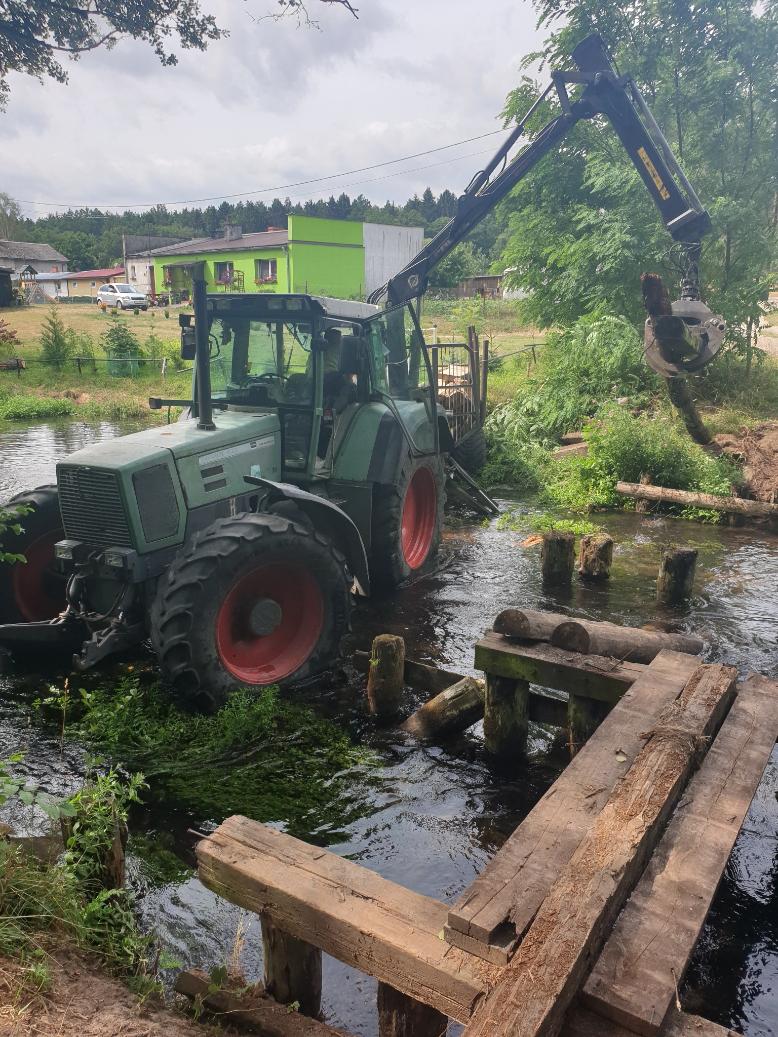 Zielony traktor Fendt z żurawiem w korycie rzeki, podnosi kawałek drewna, w tle budynki mieszkalne i zieleń.