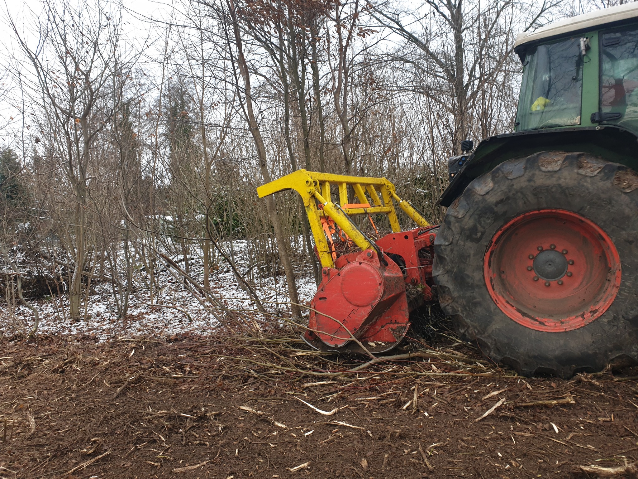 Czerwono-żółty mulczer zamontowany na traktorze, rozdrabniający gałęzie na skraju lasu w pochmurny dzień. Ziemia pokryta resztkami drewna i niewielką ilością śniegu.