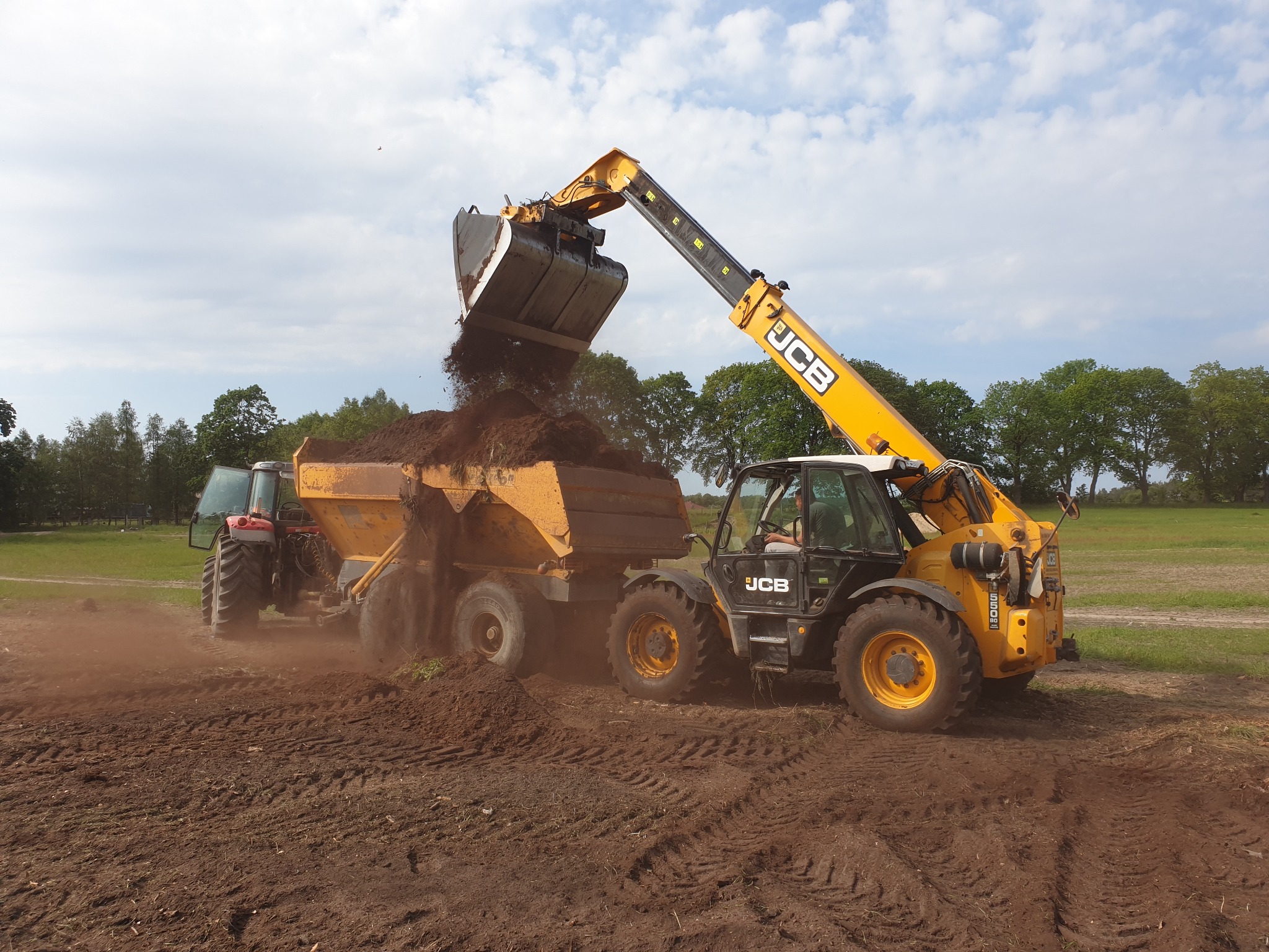 Żółty teleskopowy ładowacz JCB z łyżką wysypuje ziemię do przyczepy ciągniętej przez czerwony traktor na polu z przygotowaną glebą. Widoczne ślady opon na ziemi i drzewa w tle.