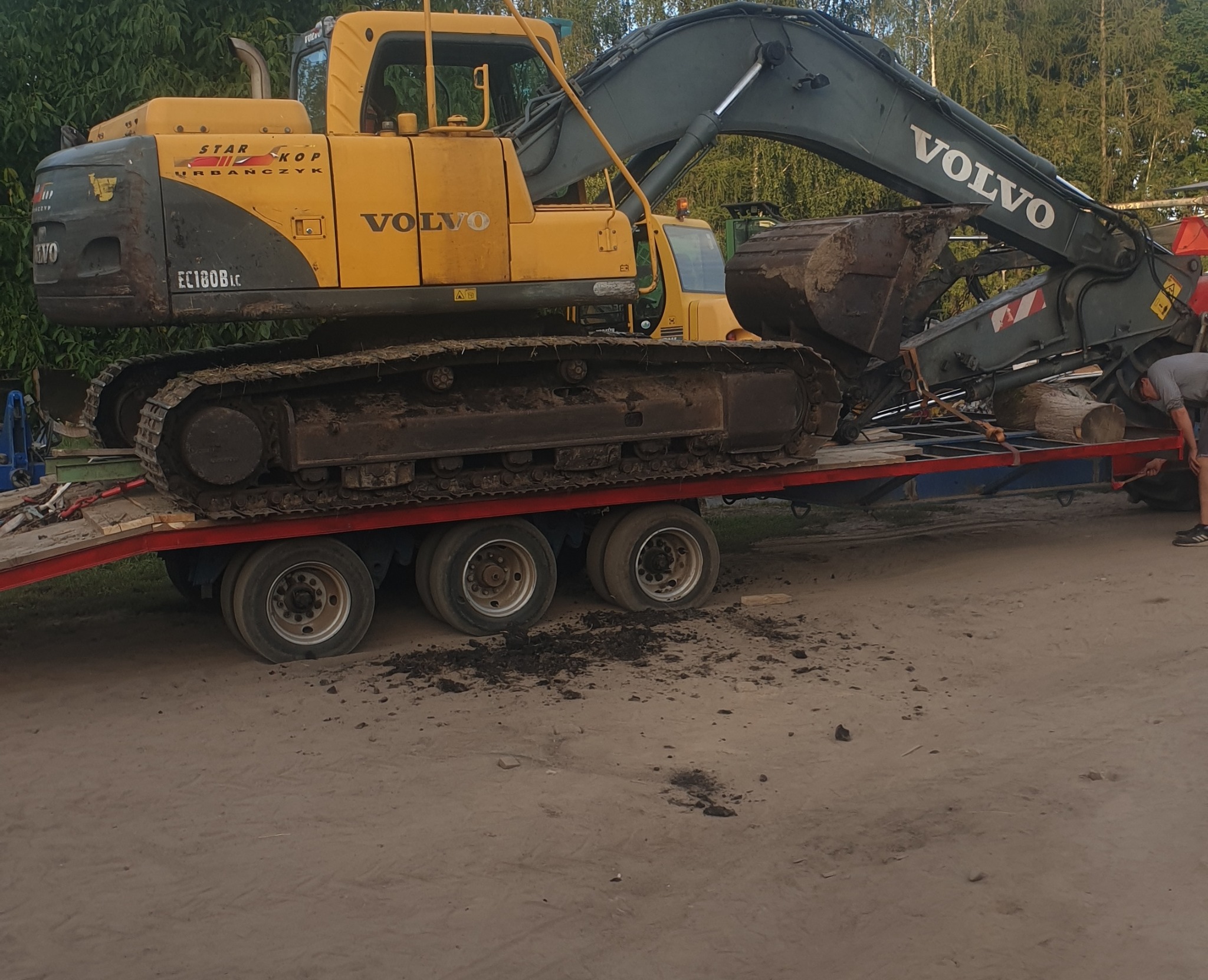 Żółto-szara koparka gąsienicowa Volvo EC180BLC, transportowana na czerwonej lawecie z trzema osiami, widoczny operator przy zabezpieczaniu ładunku.