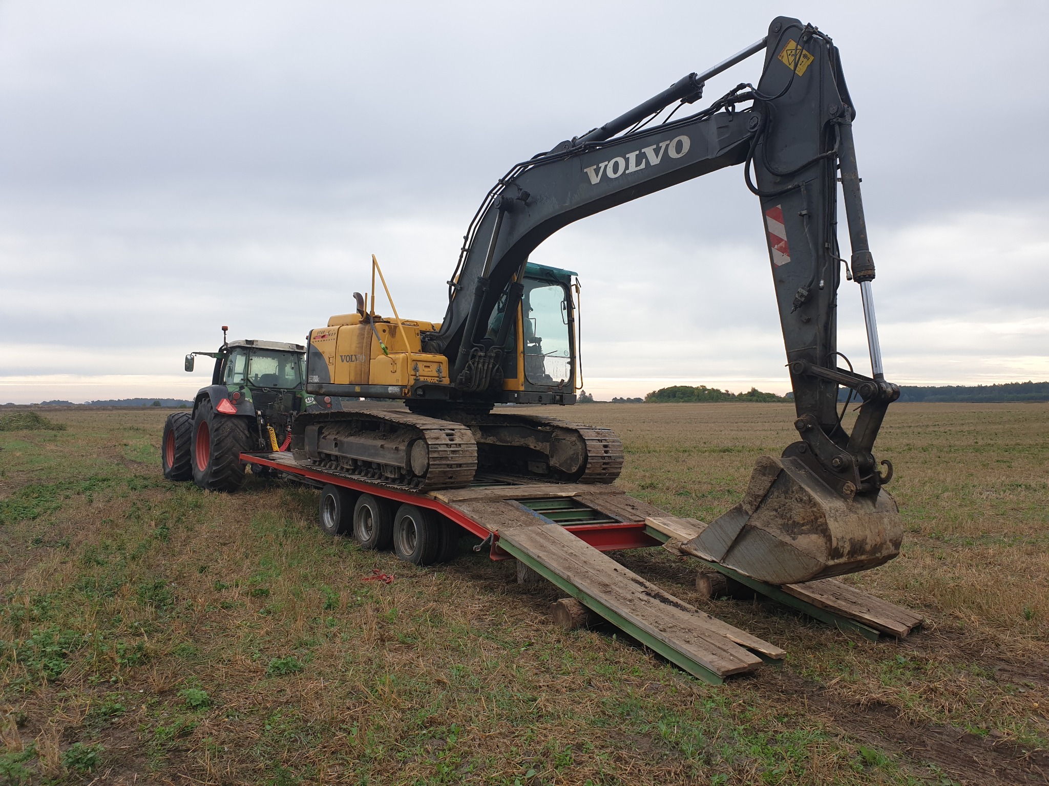 Żółto-czarna koparka Volvo na lawecie ciągniętej przez traktor na polu, pochmurne niebo w tle.