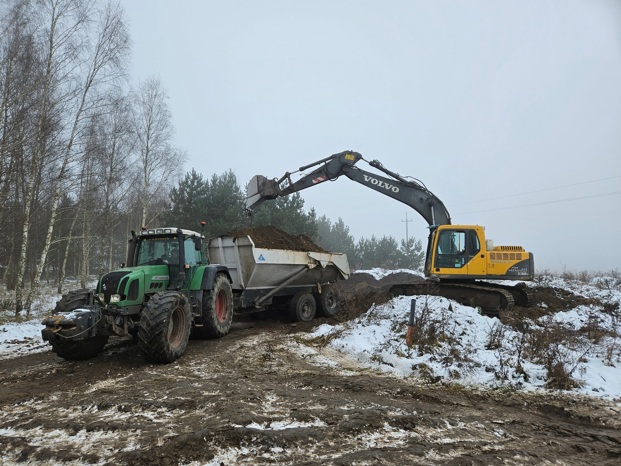 Żółta koparka Volvo nabiera ziemię i ładuje ją do przyczepy ciągnika Fendt na błotnistym, częściowo ośnieżonym terenie, drzewa w tle.