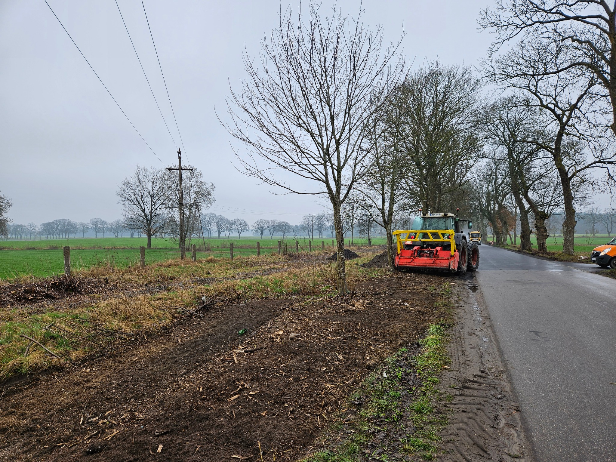 Tractor with red mulcher unit parked on the side of a wet road next to a freshly mulched area with a newly planted tree sapling; power lines and a field are visible in the background.