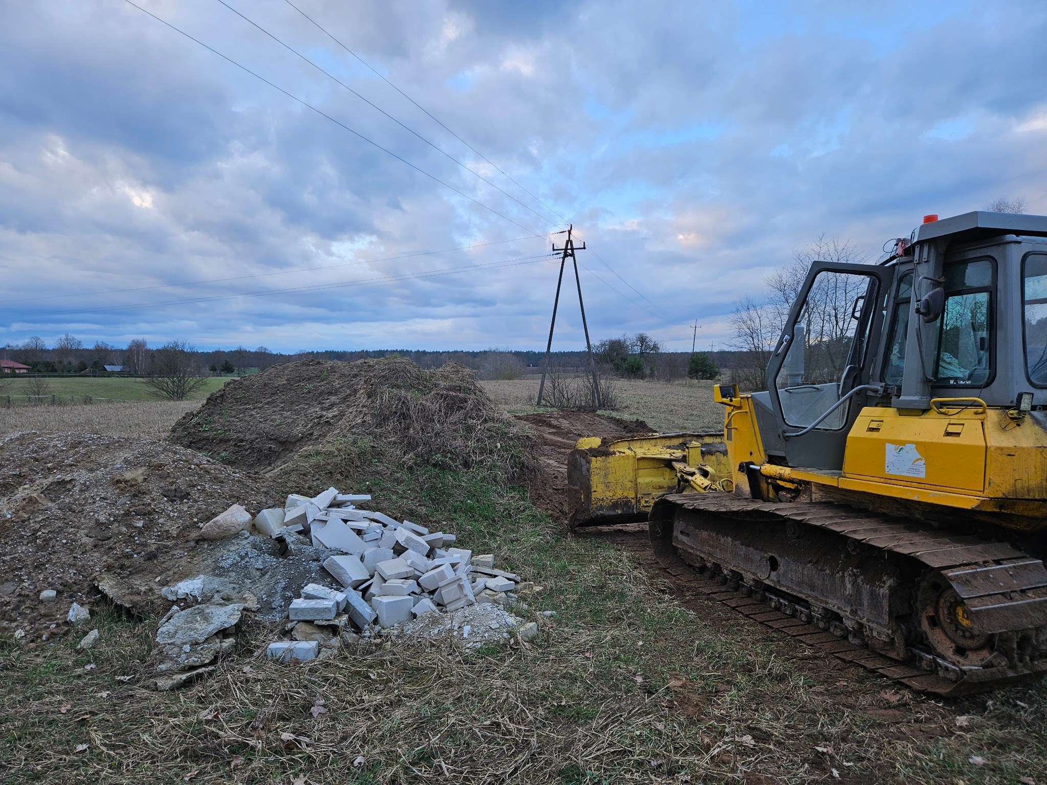 Żółty spychacz pracujący na terenie w Wałczu, obok sterta gruzu i ziemi, w tle słupy wysokiego napięcia i pochmurne niebo.