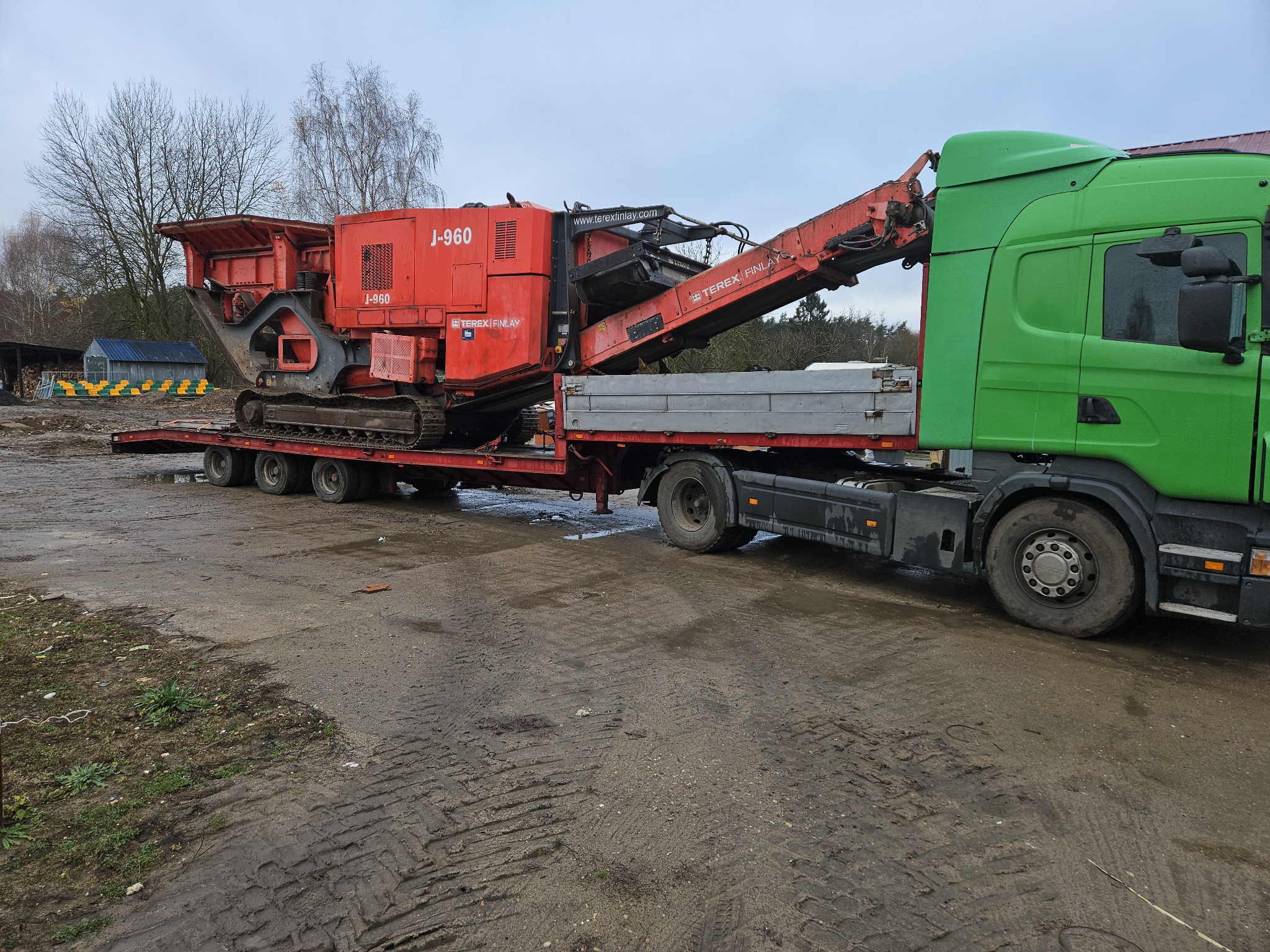 Czerwona kruszarka J-960 Terex Finlay na naczepie ciężarówki z zieloną kabiną, na błotnistym placu. Widoczne drzewa w tle.