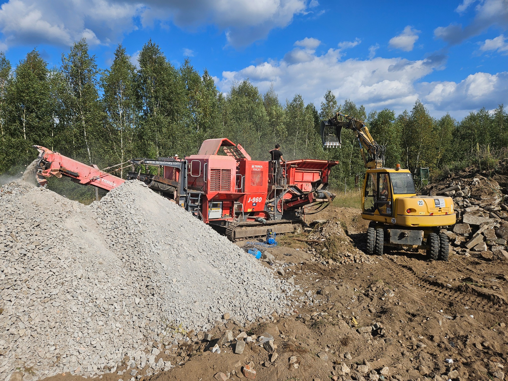 Czerwona kruszarka Terex Finlay J-960 i żółta koparka na placu budowy z hałdą gruzu na pierwszym planie, w tle drzewa i błękitne niebo z chmurami.
