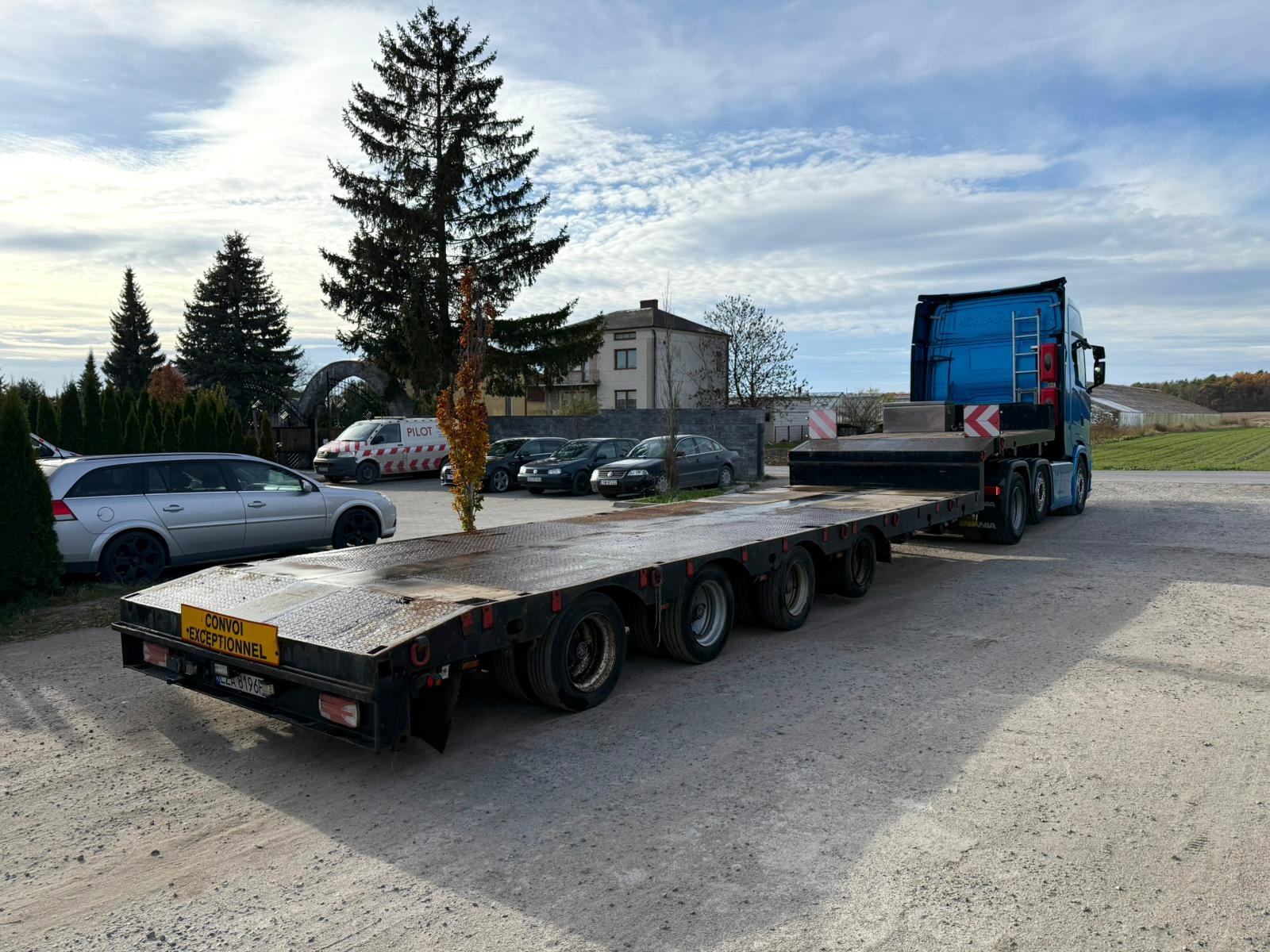 Niebieska ciężarówka z naczepą niskopodwoziową na placu, w tle budynek i drzewa. Tablica 'Convoi Exceptionnel' na naczepie. Transport ponadgabarytowy.