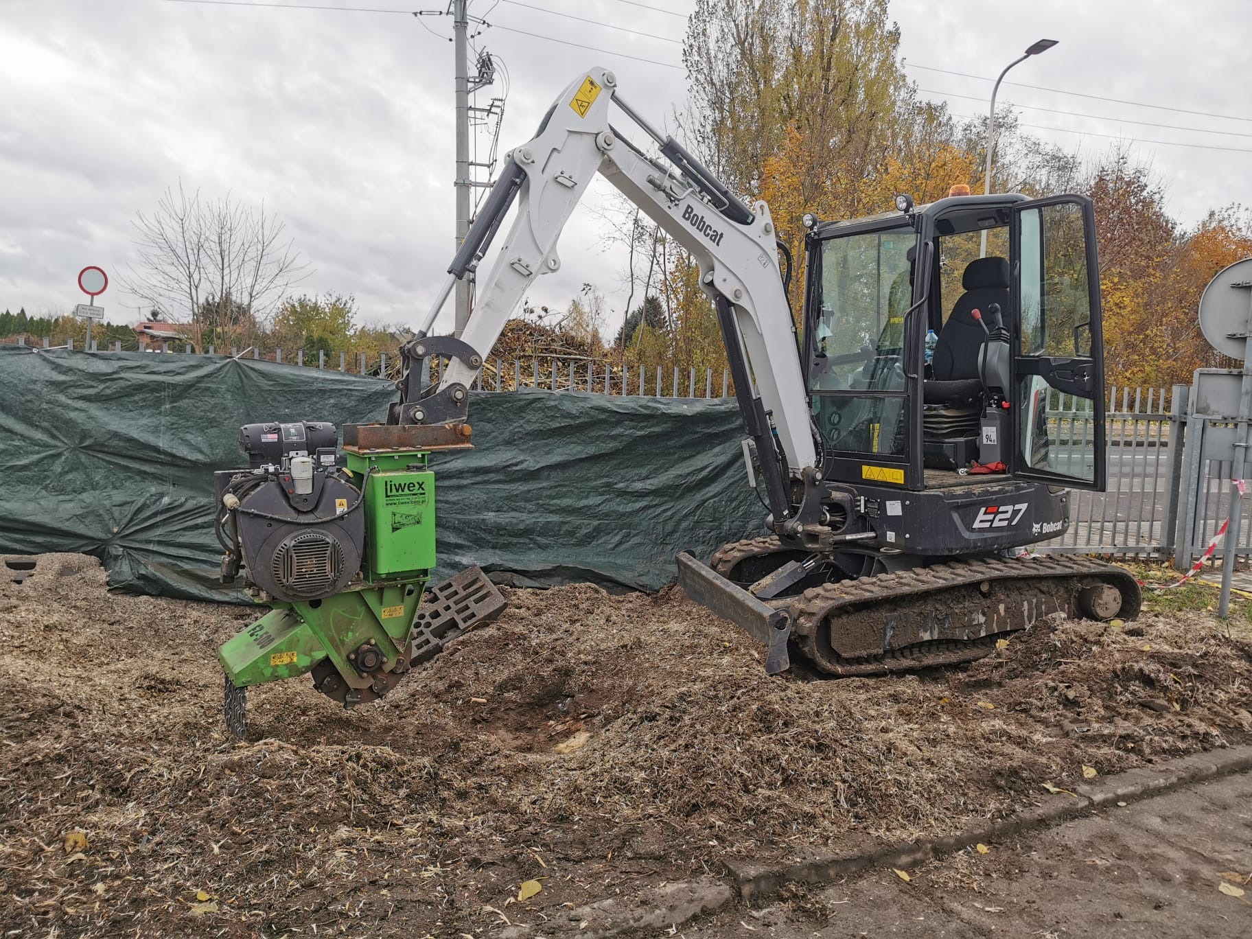 Mini koparka Bobcat E27 z frezarką do pni Iwex, usuwająca pień drzewa. Wokół rozsypane wióry, w tle zielona plandeka i jesienne drzewa.