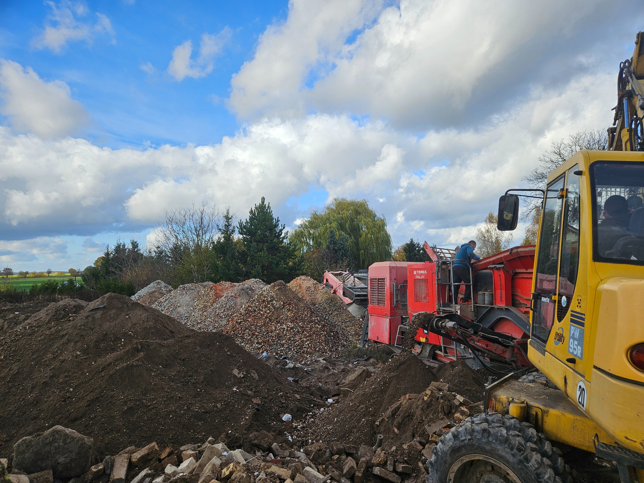 Żółta koparka i czerwona kruszarka do gruzu na placu budowy, z hałdami ziemi i gruzu. Operator na maszynie. Błękitne niebo z chmurami w tle. Widok na teren po rozbiórce.